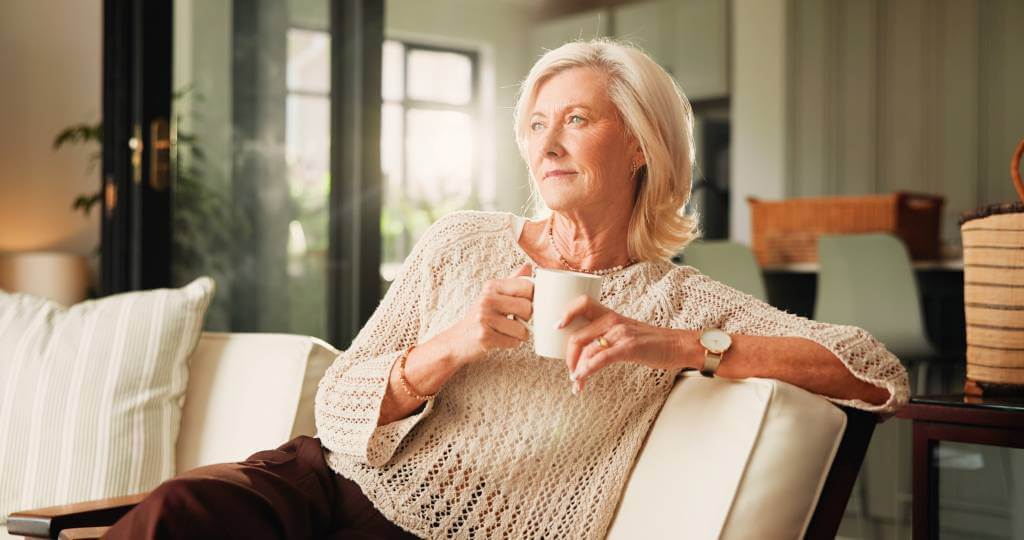 A senior woman sitting on a couch drinking coffee.