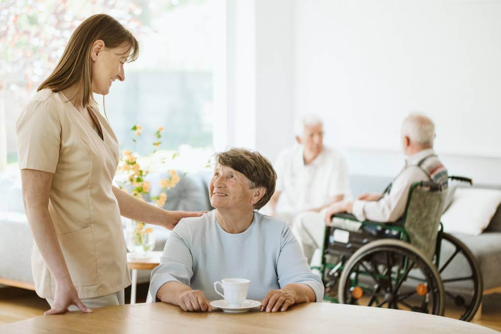 A smiling nurse helping an elderly woman in a common room of a nursing home.